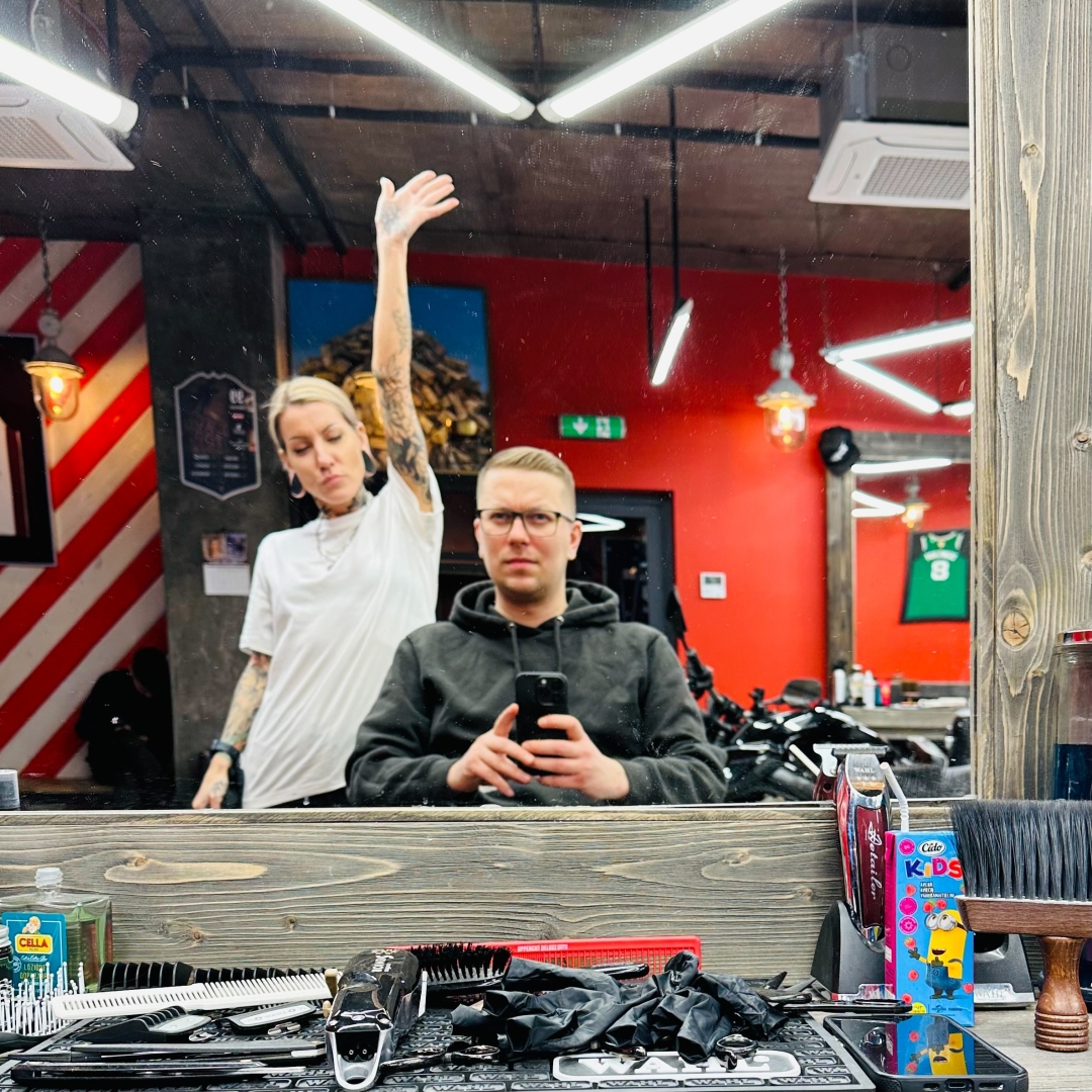 In a barbershop, showing a mirror reflection. In the mirror, a person in a black hoodie is taking a selfie while seated in a barber’s chair, while a barber in a white t-shirt with visible tattoos is standing behind them with one arm raised. The space has red walls, fluorescent lighting, and decorative elements including red and white striped panels and a basketball jersey mounted on the wall. In the foreground are various barbering tools and supplies arranged on a wooden counter, including combs, brushes, and hair products.